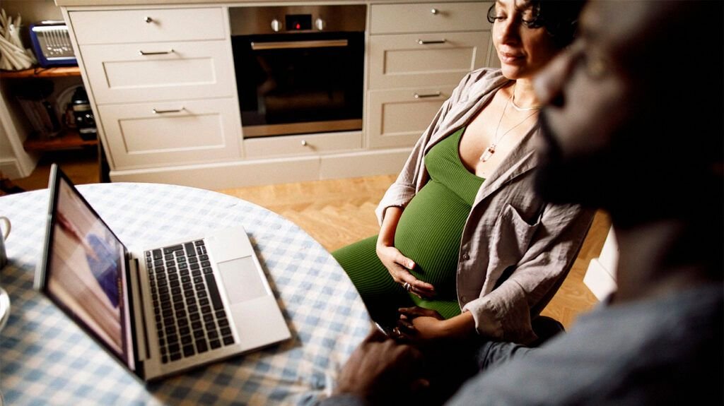 Une femme enceinte et un homme assis à une table pour un appel vidéo avec un médecin dans leur cuisine