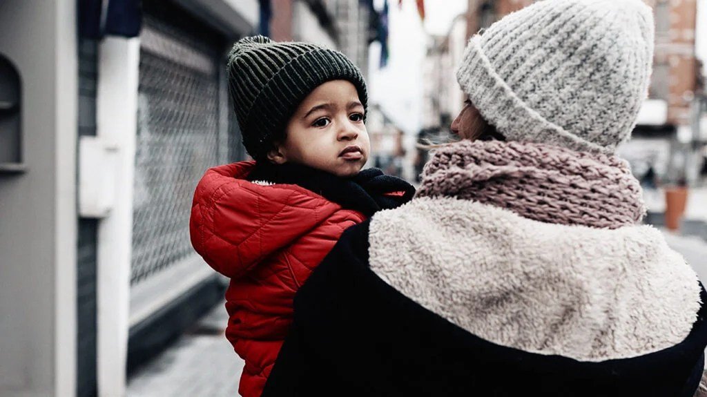 Vista posteriore della mamma che cammina per strada portando il suo bambino piccolo in braccio - Luce del giorno invernale